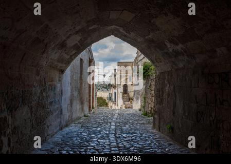 Ein bezaubernder Schuss durch einen Steinbogentunnel, der eine historische Gasse im Sassi di Matera umrahmt. Die kopfsteingepflasterte Straße führt in Richtung einer tausendjährigen Limone Stockfoto