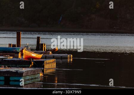 Ein kleines orangenes Ruderboot sitzt auf einem hölzernen Dock, beleuchtet von der untergehenden Sonne, mit Rudern im Inneren, bereit für die nächste Fahrt auf dem ruhigen See Stockfoto
