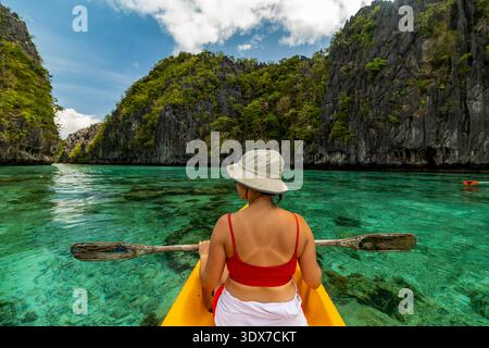 Eine Frau mit rotem Dach und Sonnenhut paddelt mit einem gelben Kajak durch klares türkisfarbenes Wasser, umgeben von felsigen, mit Bäumen bedeckten Klippen. Stockfoto