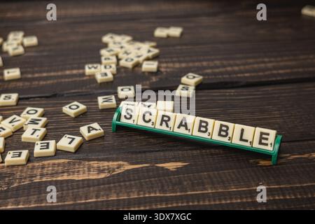 Die Fliesen sind auf einem Holztisch mit einem Scrabble-Halter verstreut, auf dem das Wort „SCRABBLE“ steht. Die Menschen genießen Spiele am National Scrabble Day zusammen. Stockfoto