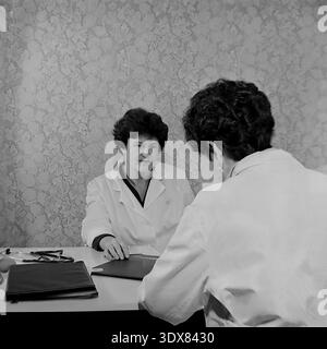 Vintage Schwarz-weiß Archivfoto, das zwei weibliche medizinische Fachkräfte in klassischen weißen Labormänteln mit einer ernsthaften professionellen Beratung einfängt. Sie sitzen einander gegenüber an einem Schreibtisch in einem Büro im Stadtkrankenhaus Nr. 2 in Sloviansk, ukrainische SSR während der 1980er Jahre Ein Stethoskop und schwarze Mappen liegen auf dem Tisch vor einem Hintergrund mit typisch sowjetisch gemusterter Blumentapete. Authentisches historisches Bild, das den Arbeitsalltag, das Gesundheitssystem und das engagierte medizinische Personal während der Sowjetzeit im friedlichen Donbass zeigt. Stockfoto