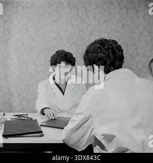 Vintage Schwarz-weiß Archivfoto, das zwei weibliche medizinische Fachkräfte in klassischen weißen Labormänteln mit einer ernsthaften professionellen Beratung einfängt. Sie sitzen einander gegenüber an einem Schreibtisch in einem Büro im Stadtkrankenhaus Nr. 2 in Sloviansk, ukrainische SSR während der 1980er Jahre Ein Stethoskop und schwarze Mappen liegen auf dem Tisch vor einem Hintergrund mit typisch sowjetisch gemusterter Blumentapete. Authentisches historisches Bild, das den Arbeitsalltag, das Gesundheitssystem und das engagierte medizinische Personal während der Sowjetzeit im friedlichen Donbass zeigt. Stockfoto