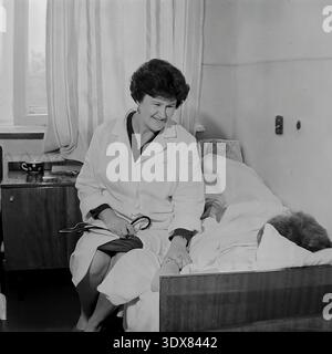 Vintage-Schwarzweiß-Archivfoto, das eine fürsorgliche Ärztin in einem klassischen weißen Labormantel am Rand eines Bettes zeigt, um einen Patienten zu beruhigen. Sie hält die Hand des Patienten und hält ein Stethoskop in einer typischen Station des Stadtkrankenhauses Nr. 2 in Sloviansk, ukrainische SSR während der 1980er Jahre Ein Nachttisch mit einem Becher mit Punktmuster steht am Fenster. Dieses authentische Bild zeigt die mitfühlende Art und Weise am Krankenbett, den täglichen Gesundheitsalltag und das engagierte medizinische Personal während der Sowjetzeit im friedlichen Donbass. Stockfoto