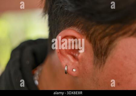 Nahaufnahme des Ohres des jungen Mannes mit zwei Lappenpiercings, kleinem silbernen Korb und Ohrstecker. Stilvolles und modernes Detail für eine Person mit dunklem Haar in lässiger Ou Stockfoto