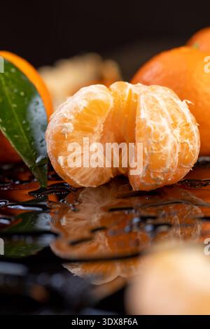 Reife Orangen und saftige und geschälte Mandarinen liegen auf einer glänzend schwarzen Oberfläche mit klaren Wassertropfen, frische weiche und süße Mandarinen auf einem schwarzen Glas c Stockfoto