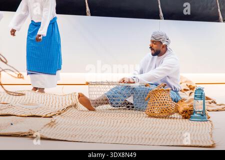 6. Dezember 2025, Abu Dhabi, VAE: Arabischer Mann in traditioneller Kleidung webt eine Fischfalle auf einer Matte und singt während einer traditionellen Vorstellung Stockfoto