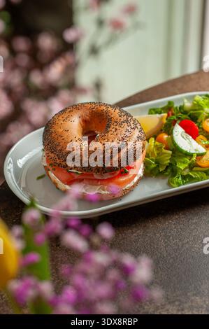 Gerösteter Mohnbagel gefüllt mit Frischkäse und Räucherlachs, serviert mit einem frischen grünen Salat aus Salat, Kirschtomaten und Gurke. Stockfoto