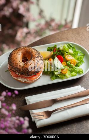 Gerösteter Mohnbagel gefüllt mit Frischkäse und Räucherlachs, serviert mit einem frischen grünen Salat aus Salat, Kirschtomaten und Gurke. Stockfoto