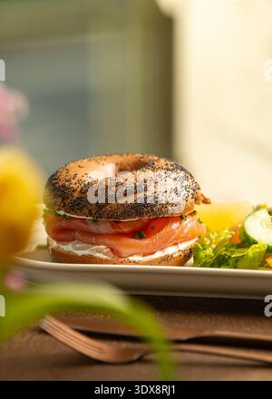 Gerösteter Mohnbagel gefüllt mit Frischkäse und Räucherlachs, serviert mit einem frischen grünen Salat aus Salat, Kirschtomaten und Gurke. Stockfoto