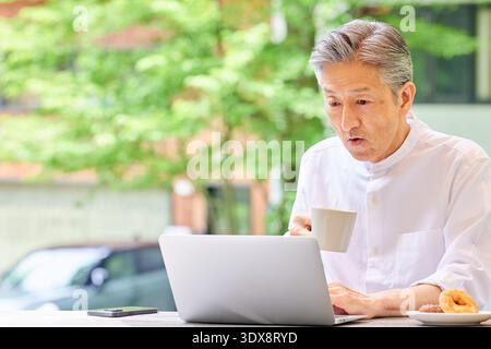 Überraschter Senior Mann, der einen Laptop benutzte und auf der Terrasse Kaffee trank Stockfoto