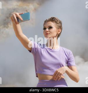 Eine selbstbewusste junge Frau mit kurzen blonden Haaren macht ein Selfie im Freien. In einem purpurfarbenen Athleisure-Set posiert sie vor einem dramatischen Hintergrund Stockfoto