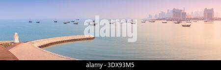 Traditionelle Dhow-Boote, die auf türkisfarbenem Wasser am Katara Beach schwimmen, mit Blick auf die Skyline von Doha und moderne Architektur während des Sonnenuntergangs Stockfoto