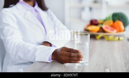 Frau im weißen Mantel hält Glas Wasser in der Nähe von frischen Früchten auf dem Tisch in einer hellen Küche Stockfoto
