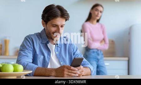Ein Mann benutzt das Smartphone, während die Frau im Hintergrund steht und die Arme in der Küche gekreuzt sind Stockfoto