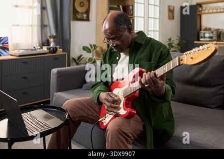 Senior Black man sitzt auf dem Sofa und spielt elektrische Gitarre, während er auf den Laptop-Bildschirm schaut, Musik zu Hause übt und sich auf das Erlernen neuer Akkorde und Techniken konzentriert Stockfoto