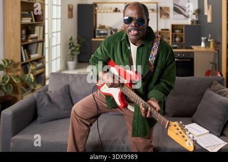 Porträt des Schwarzen Mannes mittleren Alters, der eine Sonnenbrille trägt und elektrische Gitarre spielt, im modernen Wohnzimmer steht, lächelt und in die Kamera blickt, Notenblätter auf dem Tisch sichtbar Stockfoto
