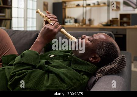 Schwarzer Mann mittleren Alters, der auf dem Sofa liegt und Holzblockflöte spielt, mit geschlossenen Augen entspannt, mit beiden Händen das Instrument hält, Küchenregale und Fenster im Hintergrund sichtbar Stockfoto