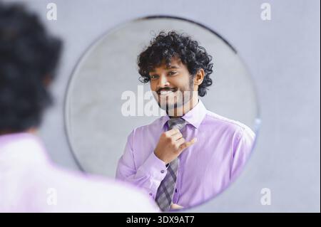 Mann, der die Krawatte vor dem Spiegel verstellt, während er ein hellviolettes Hemd trägt Stockfoto