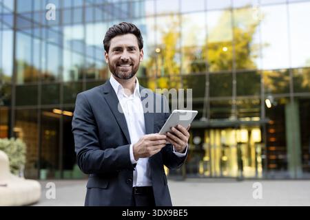 Professioneller Geschäftsmann mit Anzug und weißem Hemd lächelnd in die Kamera, während er ein digitales Tablet mit beiden Händen hält und draußen vor einem modernen Glasgebäude steht Stockfoto