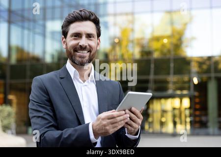 Selbstbewusster bärtiger Geschäftsmann, der vor einem modernen Bürogebäude steht, ein digitales Tablet hält und breit lächelt, steht für Erfolg, Technologie und professionelle Verbindung Stockfoto