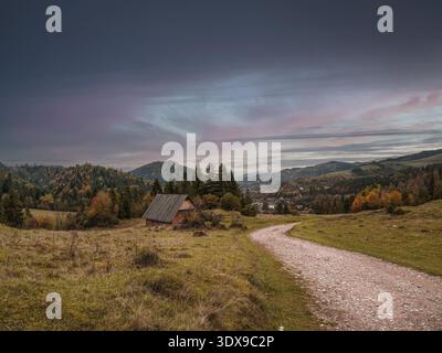 Idyllische Herbstlandschaft mit einer rustikalen Hütte inmitten von sanften Hügeln, lebhaftem Laub und einem gewundenen Pfad unter einem dramatischen, farbenfrohen Himmel. Perfekt für Stockfoto