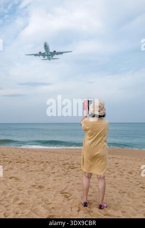 Alleinreisen im Alter Graureisen oder unheimlichen Tourismus - eine ältere aktive Frau fotografiert ein tief fliegendes Flugzeug am frühen Morgen bei Mai Khao Planes Stockfoto