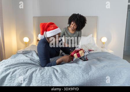 Afroamerikanisches Paar tauscht Goldgeschenk auf dem Bett ein Mann mit Weihnachtsmütze und Smartphone daneben Stockfoto