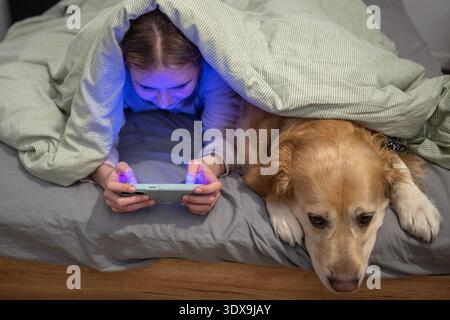 Teen Girl Spielt Mit Golden Retriever Unter Decke Und Sieht Sich Vor Dem Schlafen Das Smartphone Im Schlafzimmer An Stockfoto