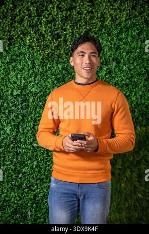 Erwachsener asiatischer Mann, der vor einer grünen Hecke steht und ein Telefon hält, mit einer orangen Sweatshirt-Jeans. Porträt, Gerät, Studio, Laub, Hintergrund, entspannt, Lifestyle || Model veröffentlicht Stockfoto