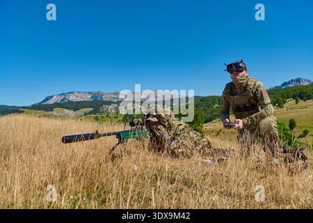 Zwei Soldaten in Tarnung führen Scharfschützentraining auf einem trockenen grasbewachsenen Feld mit Berglandschaft durch; ein vorliegender Scharfschütze und ein stehender Spotter, der einen Drohnencontroller während taktischer Übungen bedient. || Modell freigegeben Stockfoto