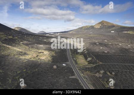 Aus der Vogelperspektive sehen Sie eine raue, vulkanische Landschaft mit Weinbergen, die in die dunkle Erde eingeätzt sind, eine Straße, die unter einem Himmel aus verstreutem Clou durch die Szene führt Stockfoto