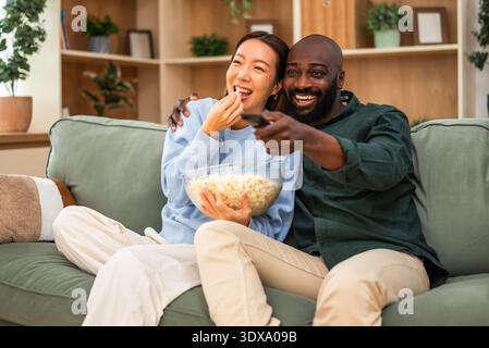 Ein fröhliches, multirassisches Paar sitzt eng zusammen auf einem grünen Sofa, lacht und umarmt, während er fernsehen und Popcorn isst. Stockfoto
