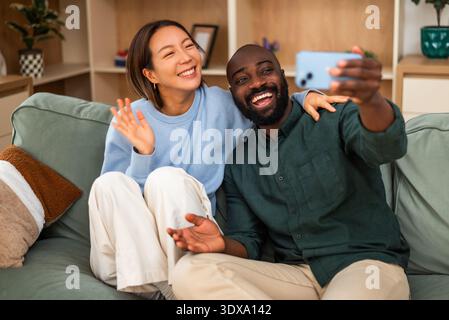 Ein fröhliches, multirassisches Paar sitzt eng zusammen auf einem grünen Sofa, lacht und umarmt in einem gemütlichen, modernen Wohnzimmer. Sie machen Selfie. Stockfoto