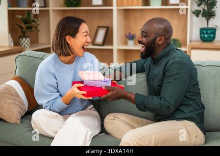 Fröhliches, multirassisches Paar hat zu Hause Spaß beim Austausch von Geschenken. Stockfoto