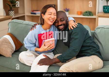 Fröhliches, multirassisches Paar hat zu Hause Spaß beim Austausch von Geschenken. Stockfoto