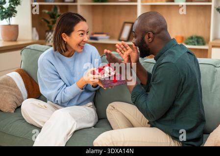 Fröhliches, multirassisches Paar hat zu Hause Spaß beim Austausch von Geschenken. Stockfoto