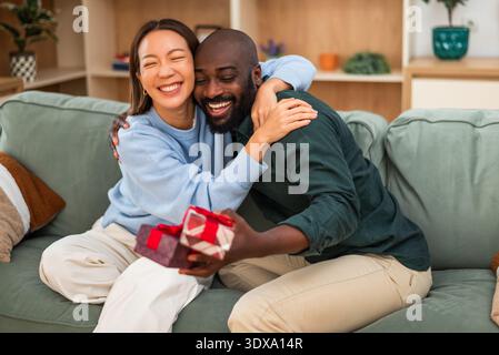 Fröhliches, multirassisches Paar, das sich zu Hause umgibt, während er Geschenke austauscht. Stockfoto