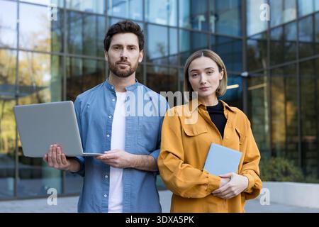 Zwei professionelle Geschäftsleute, ein Mann in der Hand eines Laptops und eine Frau in der Hand eines Tablets, stehen selbstbewusst vor einem modernen Glasbürogebäude und stehen für Teamarbeit und Unternehmenserfolg Stockfoto