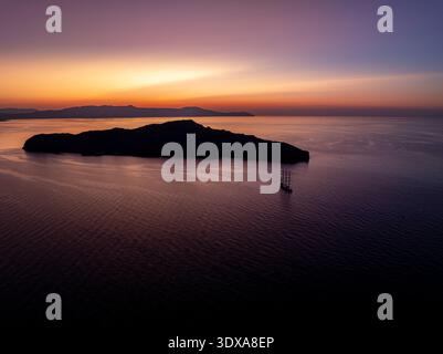 Aus der Vogelperspektive auf eine ruhige Insel, die sich vor einem pulsierenden Sonnenuntergang befindet, mit einem einsamen Segelboot, das auf den ruhigen Gewässern in Chania, Griechenland, gleitet. Stockfoto