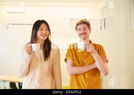 Porträt eines lächelnden Geschäftsmannes mit Kaffeetasse, der mit einer Kollegin im Büro vor einer Glaswand steht Stockfoto