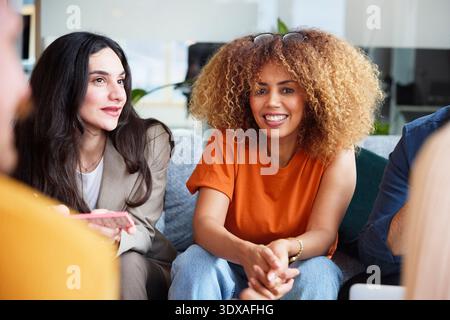 Porträt einer lächelnden, lockhaarigen Unternehmerin, die mit Kollegen im modernen Büro sitzt Stockfoto