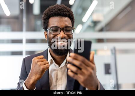 Professioneller Geschäftsmann in einem modernen Büro, der mit einem fröhlichen Lächeln und geballter Faust gute Nachrichten auf seinem Smartphone feiert und Erfolg, Leistung und selbstbewusste Begeisterung zum Ausdruck bringt Stockfoto