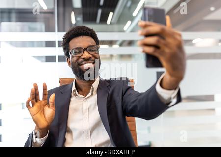 Glücklicher professioneller Mann mit Brille und Bart, der sich an einem Videoanruf beteiligt, mit der Hand winkt, während er lächelt und sein Smartphone in einem modernen Büro ansieht Stockfoto