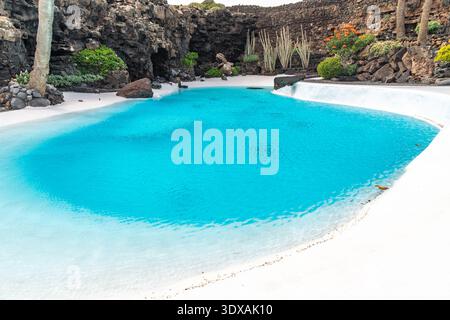 Ein ruhiger türkisfarbener Pool, eingebettet in eine vulkanische Höhle, umgeben von weißer Architektur und üppigem Grün. Die Wasseroberfläche reflektiert das Licht, Stockfoto