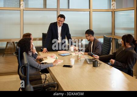 Lächelnder Manager mit Kollegen, die im Besprechungsraum um den Tisch sitzen Stockfoto