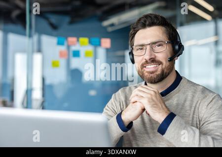 Glücklicher bärtiger Mann, der in einem modernen Büro arbeitet, ein Headset mit Mikrofon und Brille trägt, Online-Kundensupport bietet oder an einer Videokonferenz teilnimmt Stockfoto