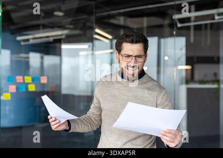 Ein gestresster junger Geschäftsmann, der frustriert ist und vor Wut schreien kann, während er Dokumente hält und schlechte Nachrichten liest, und in einem modernen Büro finanzielle Verluste und Stress erlebt Stockfoto