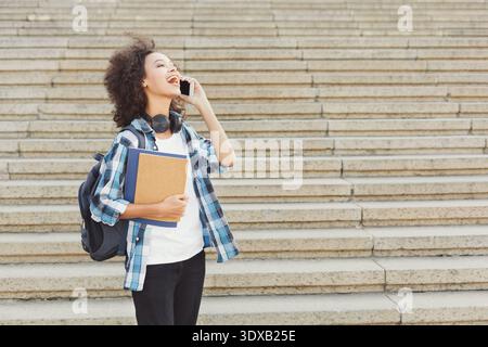 Junge Frau spricht am Telefon, während sie mit Büchern und Rucksack auf den Stufen steht Stockfoto
