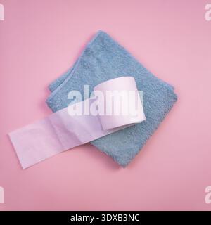 Ein hellblaues Handtuch und eine Rolle weißes Toilettenpapier sind auf rosa Hintergrund platziert. Die Szene ist einfach und minimalistisch und konzentriert sich auf das tägliche Bad Stockfoto