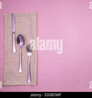 Ein Set silbernes Besteck mit Messer, Löffel und Gabel auf einer beigen Serviette vor rosa Hintergrund. Stockfoto